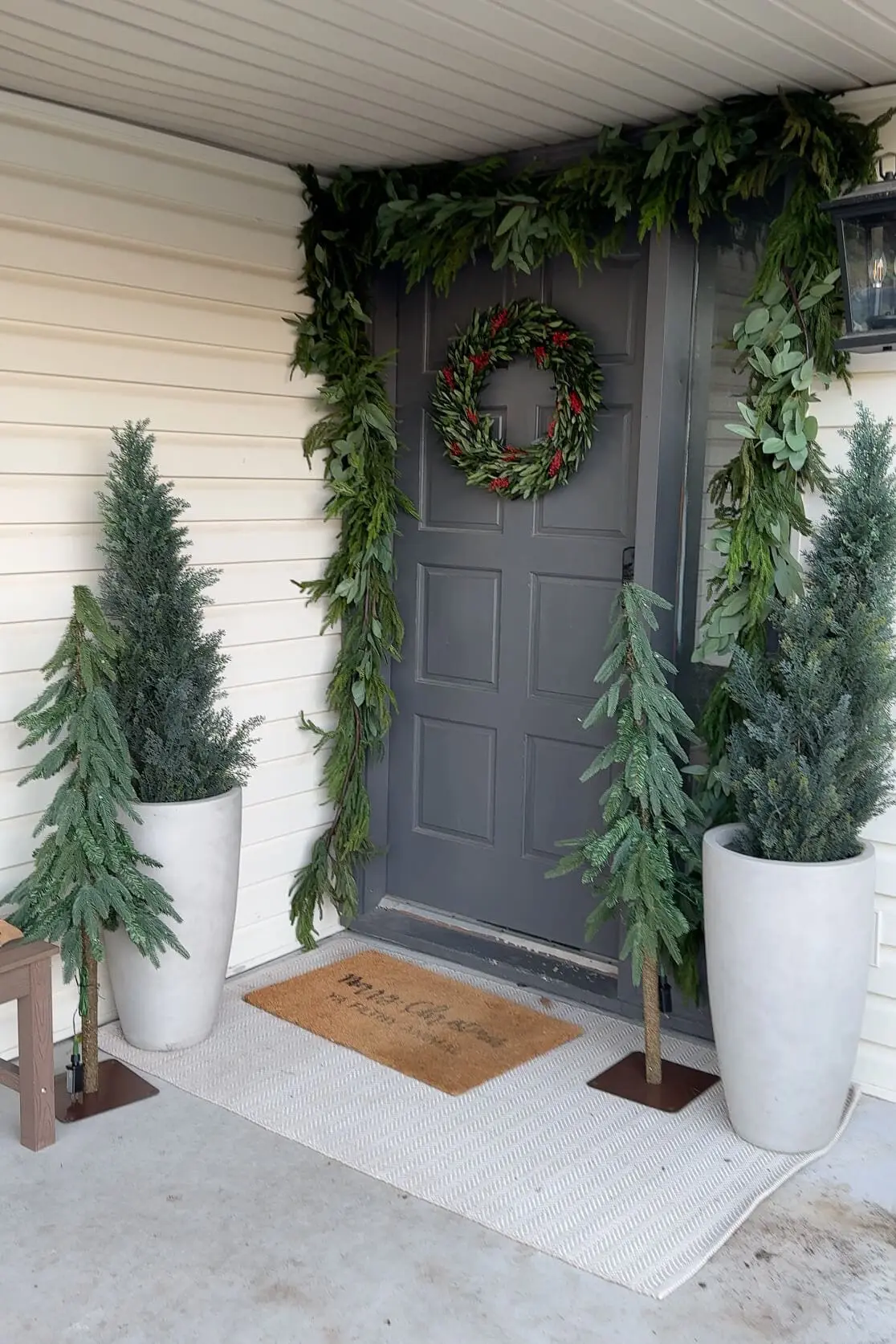 A festive front porch decorated with tall potted evergreens and small artificial Christmas trees flanking a gray door adorned with a fresh greenery wreath and garland. The setup is completed with a neutral rug and a coir doormat featuring a cheerful holiday message.